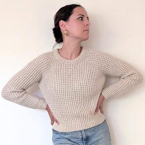 Op de afbeelding: Een vrouw draagt een crèmekleurige wafel gebreide trui en lichtblauwe jeans met patches.