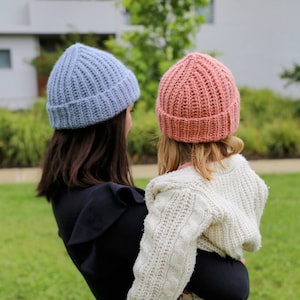 May include: A woman wearing a blue knitted beanie holds a child wearing a pink knitted beanie and a white cable knit sweater. The child is wearing brown and white leopard print leggings and blue jeans.