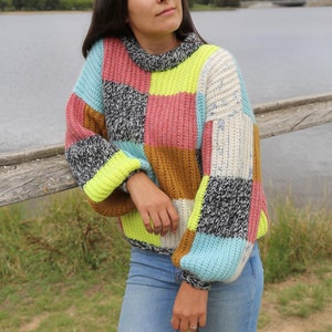 May include: A woman wearing a colourful patchwork jumper with a high neck and long sleeves. The jumper is made of different coloured squares of yarn, including pink, blue, green, white, and brown. The woman is wearing blue jeans and is standing in front of a wooden fence.