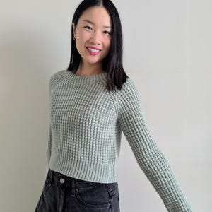 May include: A woman wearing a light green waffle knit sweater with long sleeves and a cropped hem. She is also wearing black jeans.