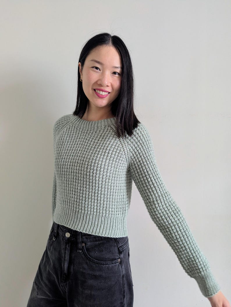 May include: A woman wearing a light green waffle knit sweater with long sleeves and a cropped hem. She is also wearing black jeans.