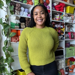 May include: A woman wearing a bright green sweater stands in front of a wall of shelves filled with colorful yarn.