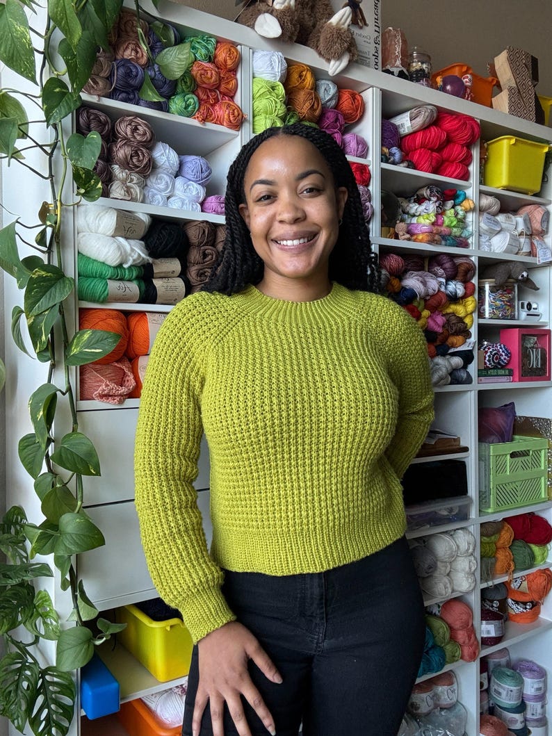 May include: A woman wearing a bright green sweater stands in front of a wall of shelves filled with colorful yarn.