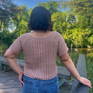 May include: A short-sleeved, light brown crochet top with a scoop neck and vertical ribbing detail. The top is paired with blue denim shorts. The background features a wooden deck, a bench, and lush green trees under a bright sky.