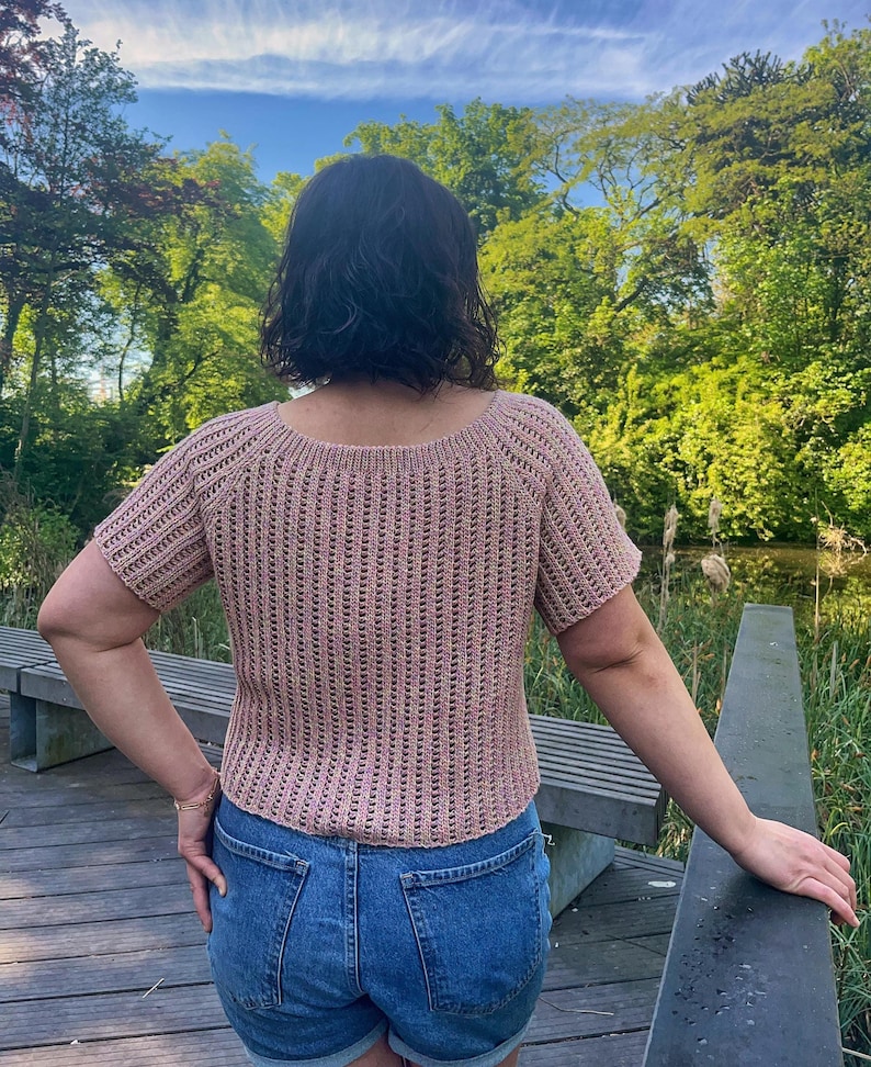 May include: A short-sleeved, light brown crochet top with a scoop neck and vertical ribbing detail. The top is paired with blue denim shorts. The background features a wooden deck, a bench, and lush green trees under a bright sky.