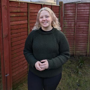 May include: A woman wearing a dark green waffle knit sweater with long sleeves and a crew neck. She is standing in front of a red wooden fence.