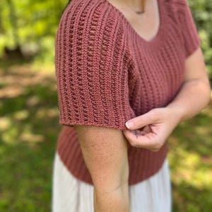 May include: A short-sleeved, brick-red knit top with a scoop neck. The sleeves have a textured, open-weave design. The top is made of a soft, breathable material, perfect for warm weather. The top is a versatile piece that can be dressed up or down.