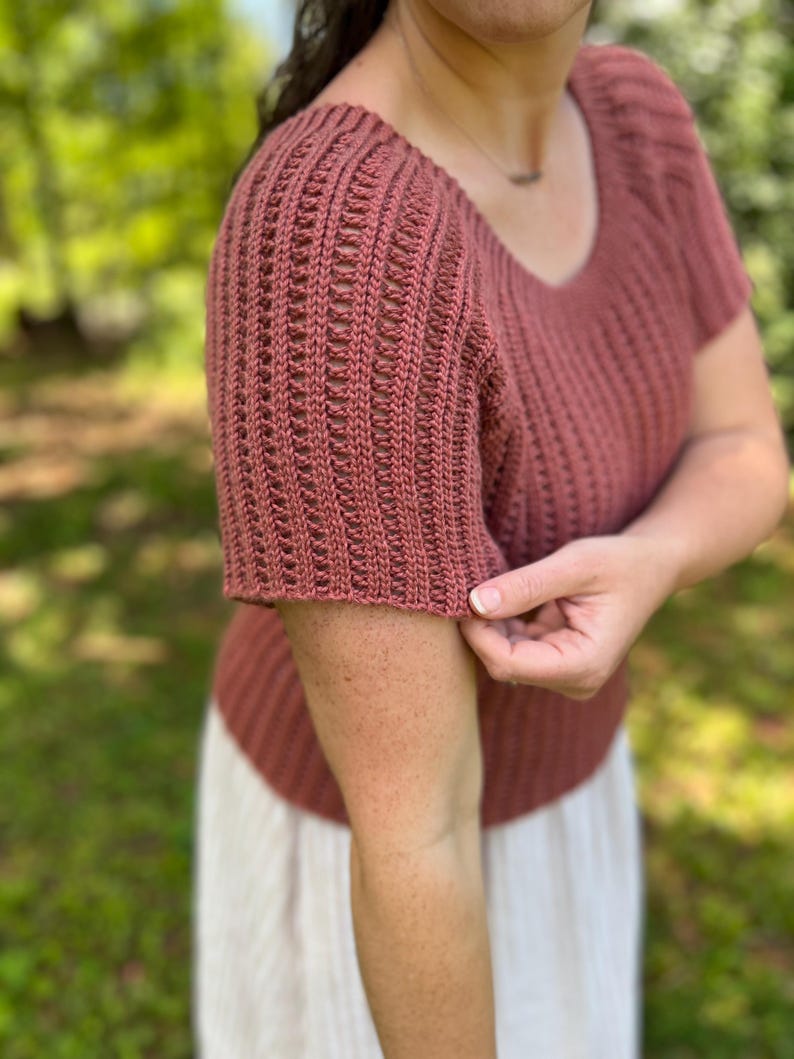 May include: A short-sleeved, brick-red knit top with a scoop neck. The sleeves have a textured, open-weave design. The top is made of a soft, breathable material, perfect for warm weather. The top is a versatile piece that can be dressed up or down.