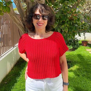 May include: A red, short-sleeved crochet top with a scoop neck. The top is paired with white jeans. The person is wearing sunglasses and a watch. The background includes a tree, a fence, and green grass.