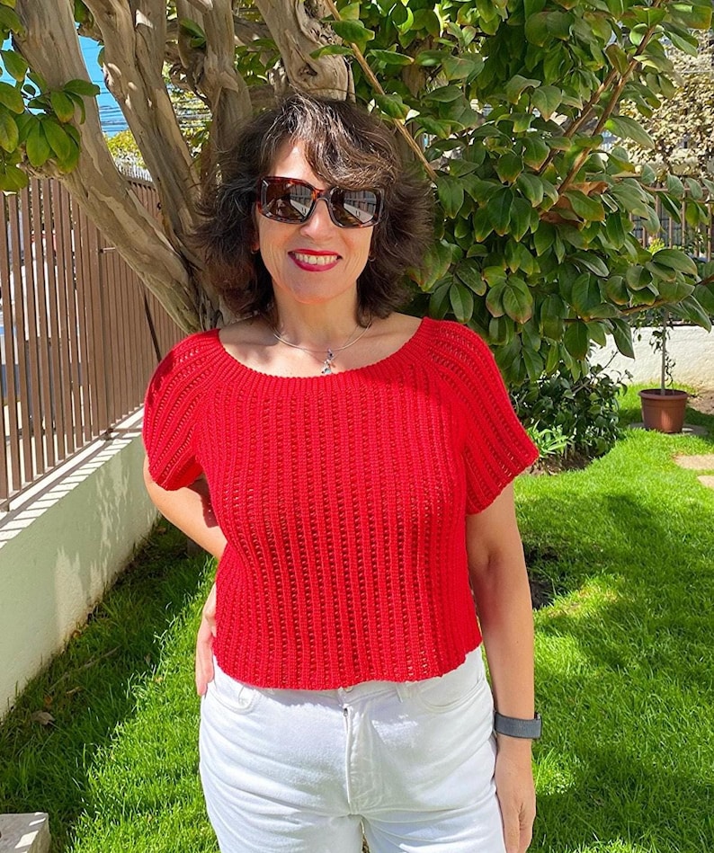 May include: A red, short-sleeved crochet top with a scoop neck. The top is paired with white jeans. The person is wearing sunglasses and a watch. The background includes a tree, a fence, and green grass.