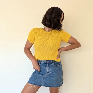 May include: A bright yellow, short-sleeved knit top paired with a denim mini skirt. The top has a ribbed texture and a crew neck. The skirt is a classic blue denim with a raw hem. The outfit is casual and stylish.