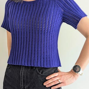 May include: A short-sleeved, royal blue crochet top with a boat neck. The top has a textured, open-weave design and is paired with black denim jeans. The overall style is casual and comfortable.