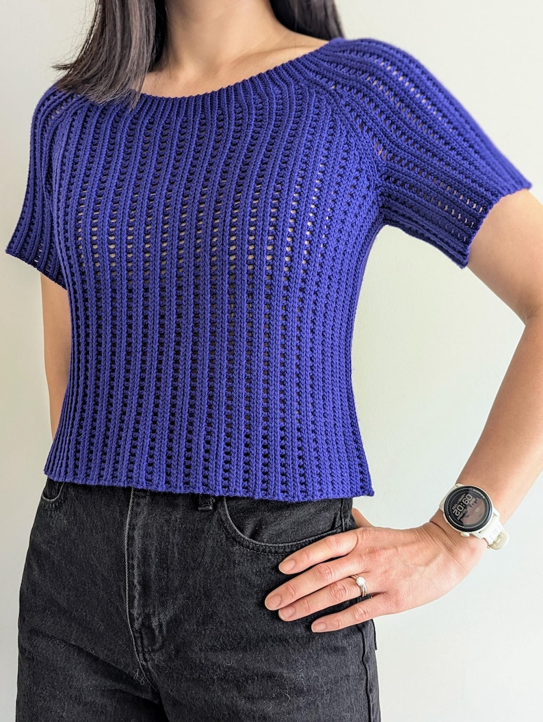 May include: A short-sleeved, royal blue crochet top with a boat neck. The top has a textured, open-weave design and is paired with black denim jeans. The overall style is casual and comfortable.