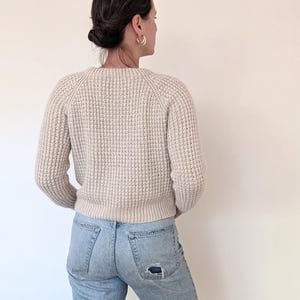 May include: A woman wearing a cream-colored knitted sweater with a waffle stitch and light blue jeans with a distressed detail on the back pocket.