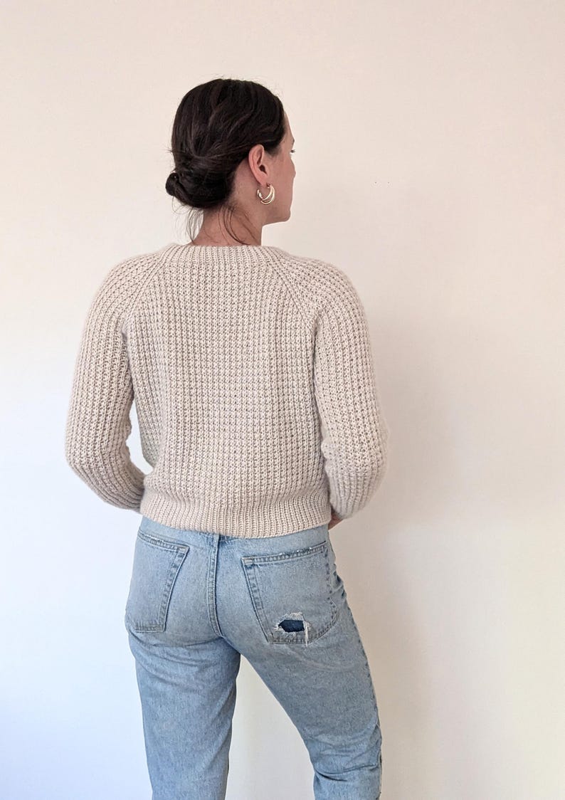 May include: A woman wearing a cream-colored knitted sweater with a waffle stitch and light blue jeans with a distressed detail on the back pocket.