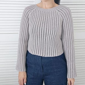 May include: A light gray, long-sleeved crochet sweater with a round neckline. The sweater has a ribbed design with vertical openwork detailing. The sleeves are slightly flared. The person is wearing blue jeans.