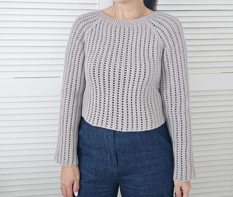 May include: A light gray, long-sleeved crochet sweater with a round neckline. The sweater has a ribbed design with vertical openwork detailing. The sleeves are slightly flared. The person is wearing blue jeans.