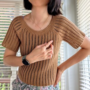 May include: A short-sleeved, brown crochet top with a scoop neckline. The open-weave design of the shirt allows for breathability. The top is a solid color and appears to be handmade. The shirt is a neutral color and can be paired with many different bottoms.