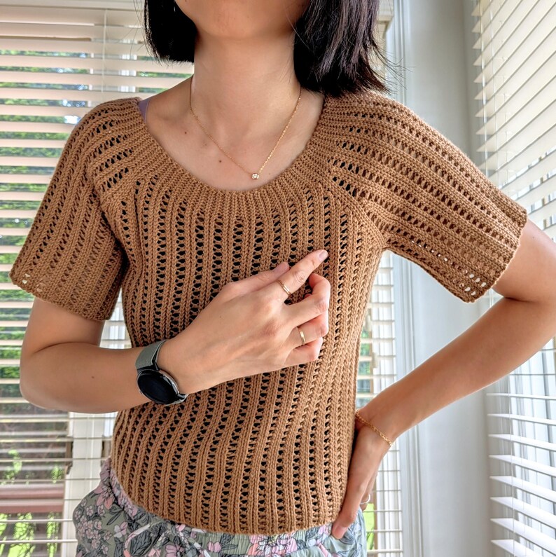 May include: A short-sleeved, brown crochet top with a scoop neckline. The open-weave design of the shirt allows for breathability. The top is a solid color and appears to be handmade. The shirt is a neutral color and can be paired with many different bottoms.