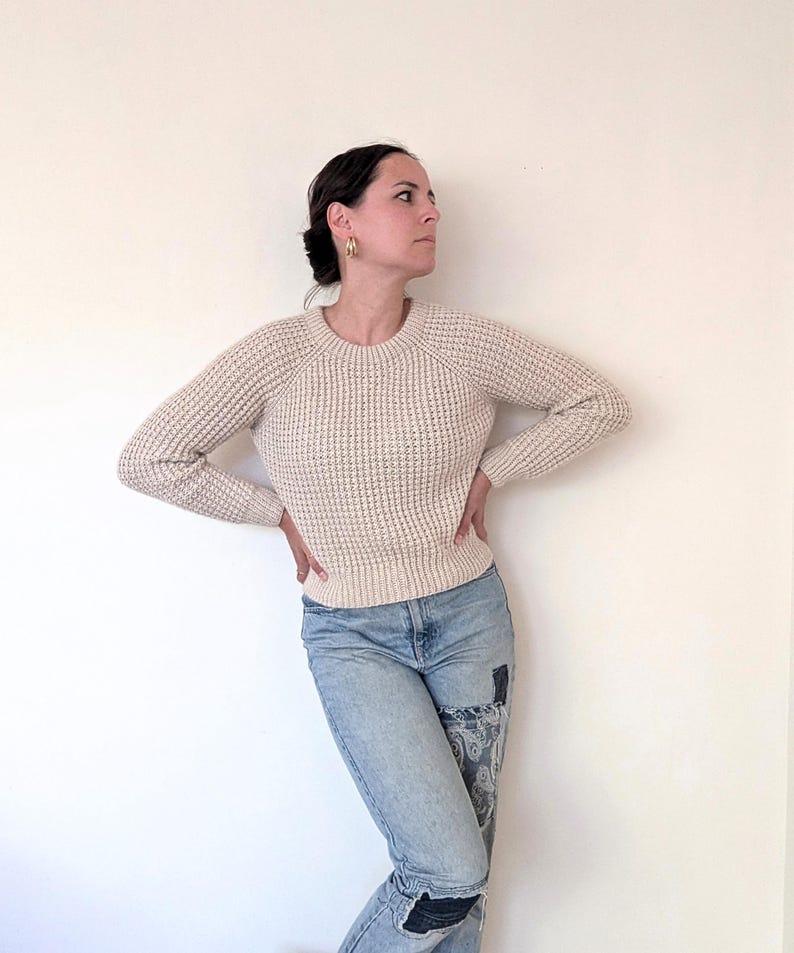 May include: A woman wearing a cream waffle knit sweater and light blue jeans with patches.