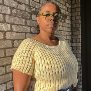 May include: A short-sleeved, yellow knit top with a scoop neckline. The top has a ribbed texture and is worn with blue jeans. The person in the image is wearing green-framed glasses.