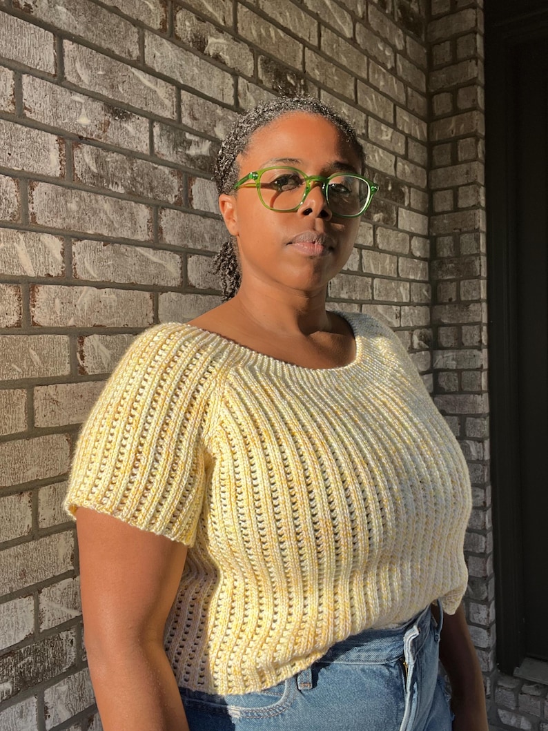 May include: A short-sleeved, yellow knit top with a scoop neckline. The top has a ribbed texture and is worn with blue jeans. The person in the image is wearing green-framed glasses.