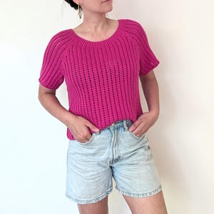 Può includere: Un top all'uncinetto magenta a maniche corte con scollo tondo. Il top presenta un motivo a coste verticali ed è abbinato a pantaloncini di jeans chiari. La persona indossa orecchini a cerchio dorati.