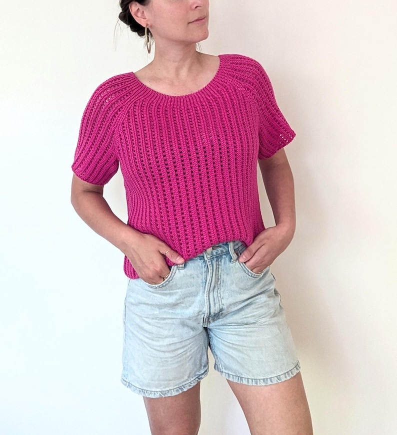 May include: A short-sleeved, magenta crochet top with a scoop neck. The top has a vertical ribbed pattern and is paired with light-wash denim shorts. The model is wearing gold hoop earrings.