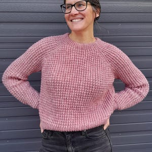 May include: A pink knitted sweater with a round neckline and long sleeves. The sweater has a waffle knit texture.