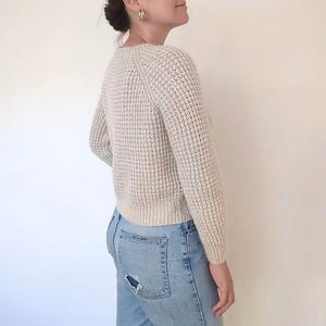 May include: A cream-colored waffle knit sweater with a round neckline and long sleeves. The sweater is cropped and has a relaxed fit.