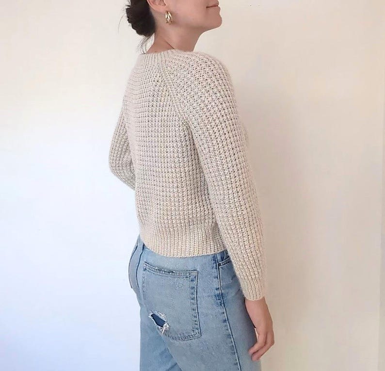 May include: A cream-colored waffle knit sweater with a round neckline and long sleeves. The sweater is cropped and has a relaxed fit.