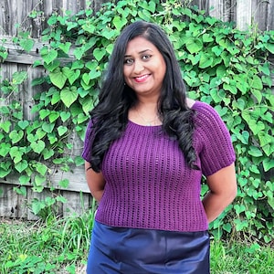 May include: A woman wearing a purple crochet top with short sleeves and a scoop neckline. The top has a ribbed texture. She is also wearing a navy blue skirt. The background includes a wooden fence and green foliage.