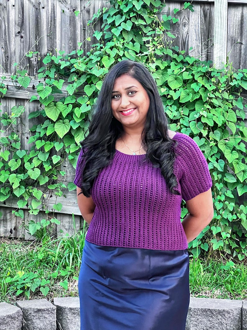 May include: A woman wearing a purple crochet top with short sleeves and a scoop neckline. The top has a ribbed texture. She is also wearing a navy blue skirt. The background includes a wooden fence and green foliage.