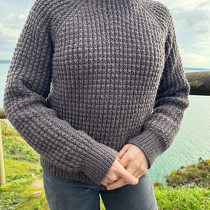 May include: A dark gray knitted sweater with a round neckline and long sleeves. The sweater has a textured waffle stitch pattern.