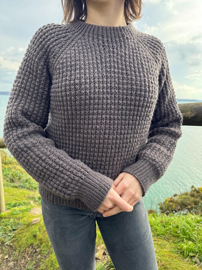 May include: A dark gray knitted sweater with a round neckline and long sleeves. The sweater has a textured waffle stitch pattern.