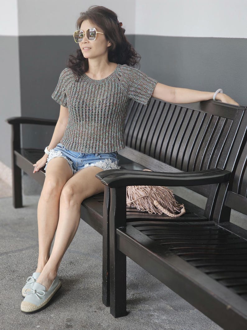 May include: A gray, short-sleeved, open-weave top. The top is paired with denim shorts and light blue shoes. The person is wearing sunglasses and sitting on a dark wooden bench. A woven bag rests on the bench.