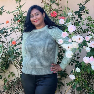 May include: A woman wearing a light green waffle knit sweater with a round neckline and long sleeves. She is standing in front of a bush with pink and white flowers.