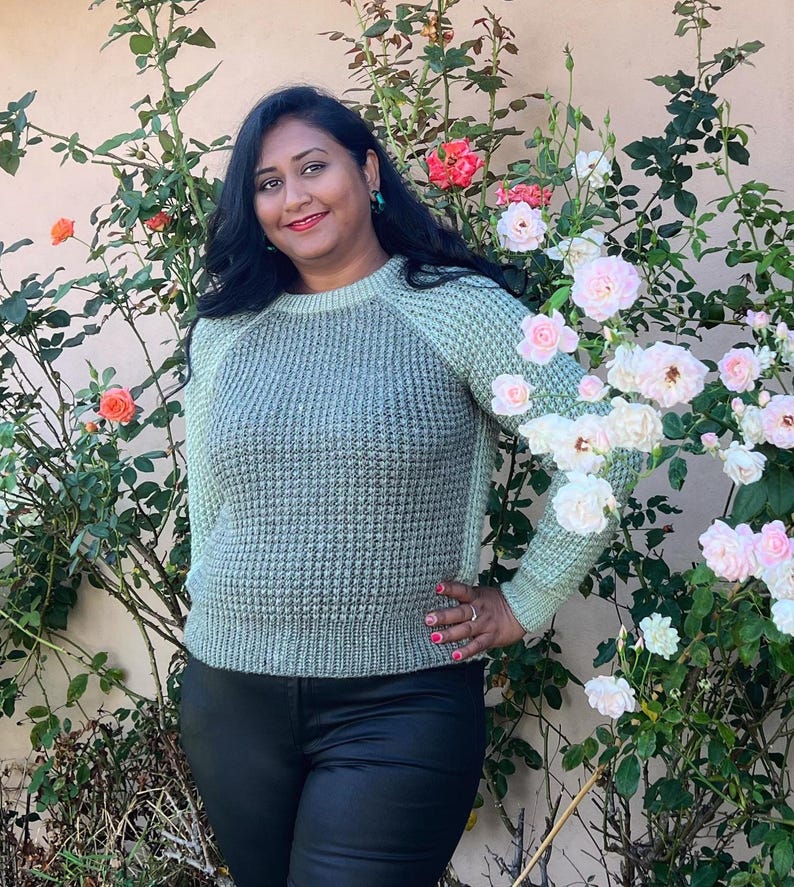 May include: A woman wearing a light green waffle knit sweater with a round neckline and long sleeves. She is standing in front of a bush with pink and white flowers.