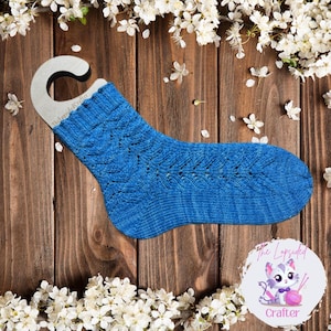 May include: A blue knitted sock with a lacy pattern, displayed on a wooden hanger against a woodgrain background with white flowers.