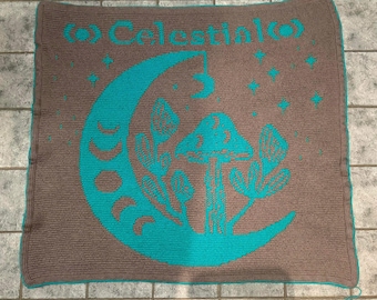 Puede incluir: Una manta de crochet con fondo marrón y un diseño de luna creciente turquesa. La luna presenta un hongo y plantas. El texto "Celestial" está escrito en turquesa en la parte superior de la manta.