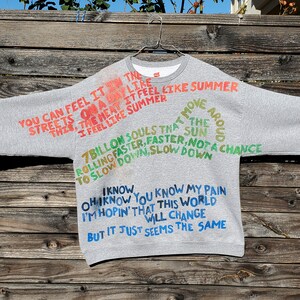 Pode incluir: Camisola de moletom cinza com texto pintado &agrave; m&atilde;o em cores. O texto diz "You can feel it in the streets on a day like this, the heat it feel like summer, I feel like summer. 7 billion souls that move around the sun rolling faster, faster, not a chance to slow down, slow down. I know, oh I know you know my pain I'm hopin' that this world will change but it just seems the same."
