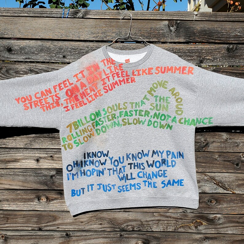 Pode incluir: Camisola de moletom cinza com texto pintado &agrave; m&atilde;o em cores. O texto diz "You can feel it in the streets on a day like this, the heat it feel like summer, I feel like summer. 7 billion souls that move around the sun rolling faster, faster, not a chance to slow down, slow down. I know, oh I know you know my pain I'm hopin' that this world will change but it just seems the same."