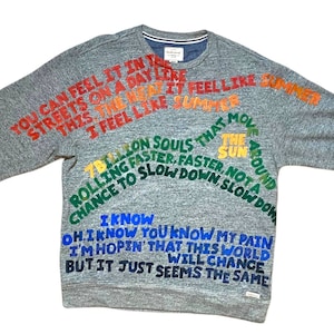 Pode incluir: Uma camisa de moletom cinza com texto colorido impresso nela. O texto diz "You can feel it in the streets on a day like this, the heat, I feel like summer, 7 billion souls that move rolling faster, faster, chance to slow down, not a slow down, I know oh, I know you know my pain, I'm hopin' that this world will change but it just seems the same".
