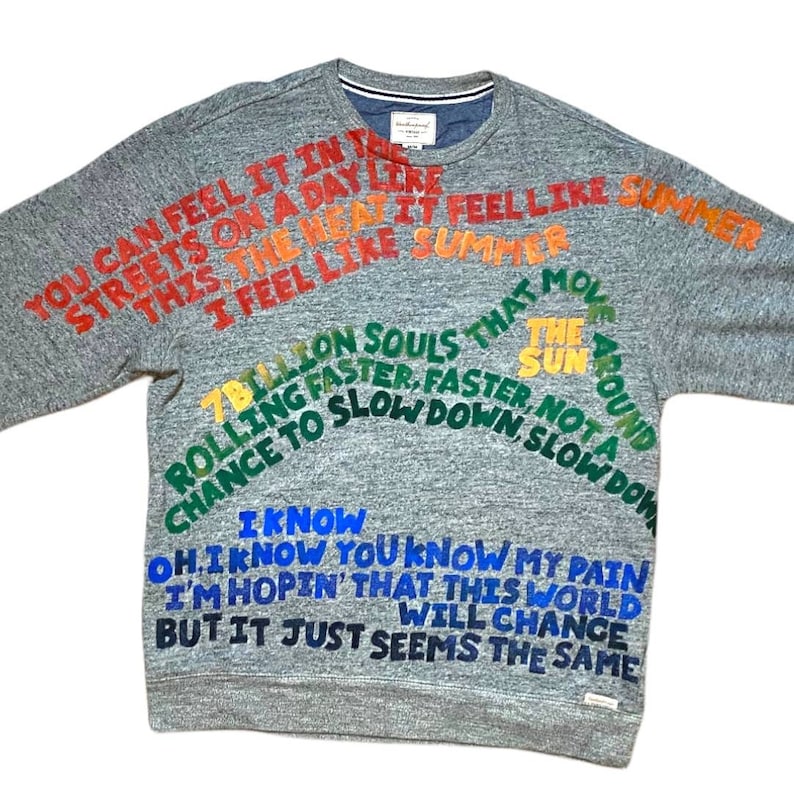 Pode incluir: Uma camisa de moletom cinza com texto colorido impresso nela. O texto diz "You can feel it in the streets on a day like this, the heat, I feel like summer, 7 billion souls that move rolling faster, faster, chance to slow down, not a slow down, I know oh, I know you know my pain, I'm hopin' that this world will change but it just seems the same".
