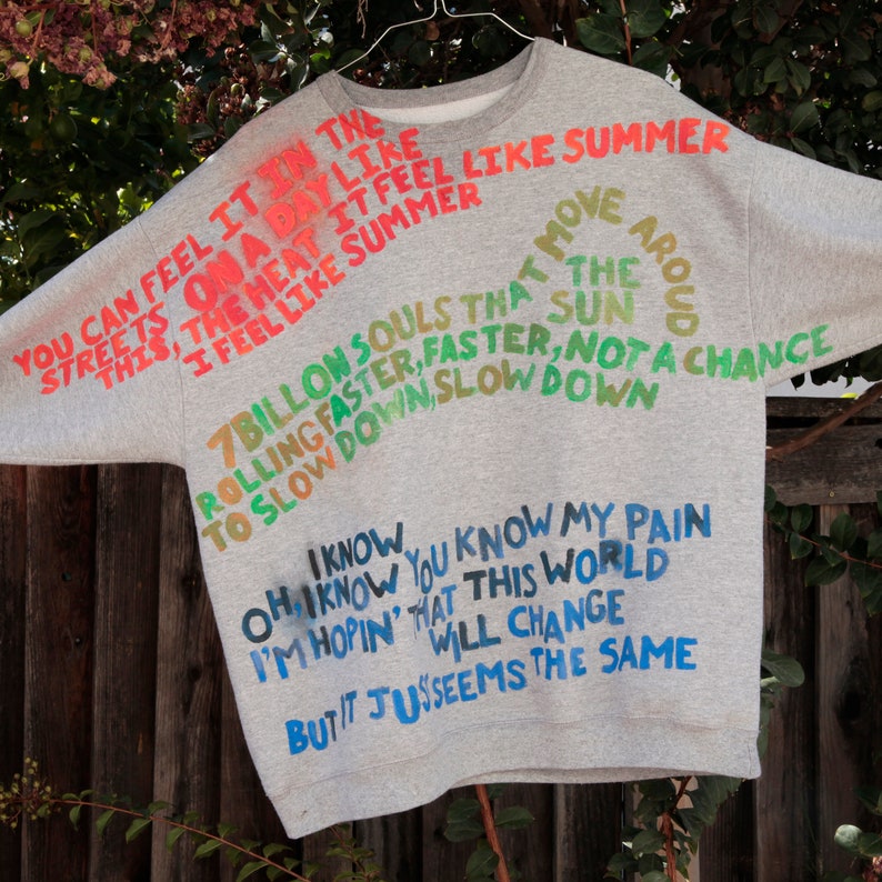 Pode incluir: Camisola de cor cinzenta com texto colorido impresso nela. O texto diz "You can feel it in the streets on a day like this, the heat it feel like summer I feel like summer move at move the sun 7 billion souls that rolling faster, faster, not a chance to slow down, slow down I know oh I know you know my pain I'm hopin' that this world will change but it jus seems the same"