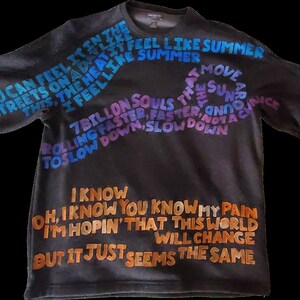 Pode incluir: Uma camisa preta de manga comprida com texto colorido estampado na frente. O texto diz "You can feel it in the streets on a day like this, the heat it feel like summer, I feel like summer, 7 billion souls that move around the sun, rolling faster, faster, and not a chance to slow down, slow down. I know, oh, I know you know my pain, I'm hopin' that this world will change, but it just seems the same."