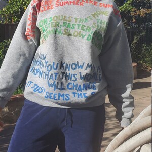 Pode incluir: Camisola de moletom cinza com texto colorido que diz "The day it feel like summer on souls that move faster, faster, and down, slow down. I know you know my heart, in that this world will change it just seems the..."