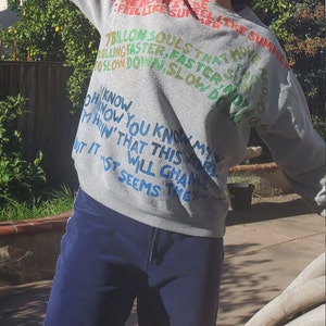Pode incluir: Camisola de moletom cinza com texto colorido que diz "You can feel the streets on fire, feel like summer, 7 billion souls that move faster, faster, slow down slow down, Oh now you know my marin' that this world will change, but it seems that..."
