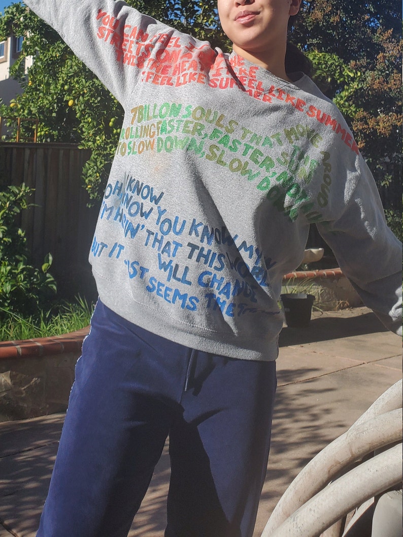 Pode incluir: Camisola de moletom cinza com texto colorido que diz "You can feel the streets on fire, feel like summer, 7 billion souls that move faster, faster, slow down slow down, Oh now you know my marin' that this world will change, but it seems that..."