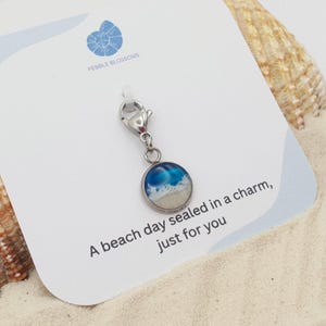 May include: A silver charm with a round pendant depicting a miniature beach scene with blue waves and sandy shore. The charm is attached to a lobster clasp. The card reads "A beach day sealed in a charm, just for you."